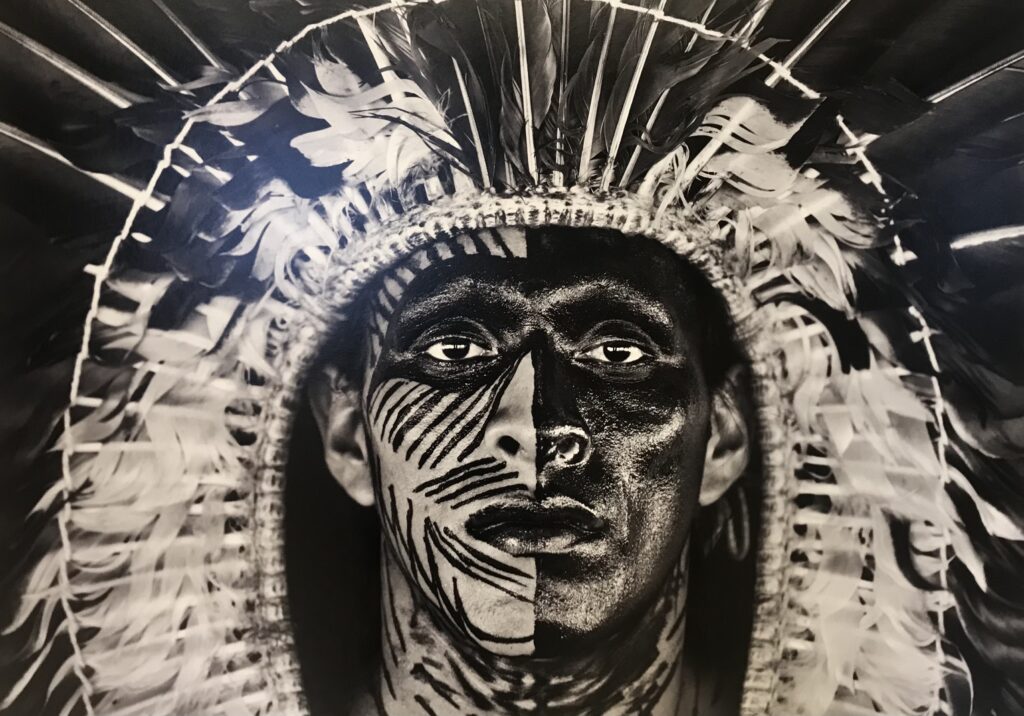 Photo en noir et blanc de Sebastiao Salgadore, représente Adao avec une coiffe en plumes d’aigle et une peinture sur le visage.